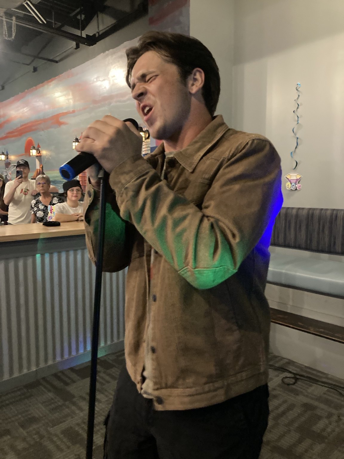 Man in a tan jacket singing into a microphone at a bar